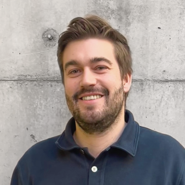 Portraitfoto von Adrian Schrödter vor einer Betonwand, er lächelt in die Kamera