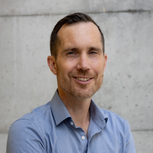 Portraitfoto von Benjamin Bartmann vor einer Betonwand, er lächelt in die Kamera